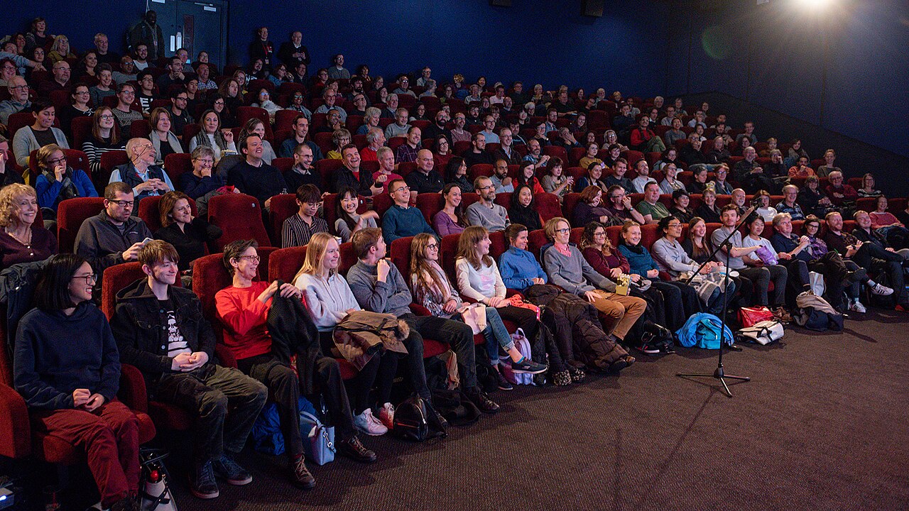 A full cinema audience seated in red velvet seats, watching the screen at a film festival.